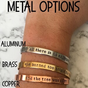 May include: Three metal bracelets with different inscriptions. The top bracelet is silver and says "after all there is no enjoyment". The middle bracelet is gold and says "she burned too bright for the". The bottom bracelet is copper and says "and the tree was happy".