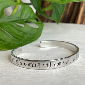 May include: A silver-toned metal cuff bracelet with an open design. The outer surface of the bracelet is engraved with the black text, "what's coming will come and we'll meet it when it does," written in a simple, clean, sans-serif font style.