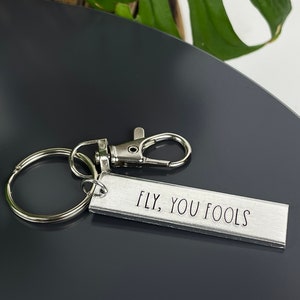 May include: Silver keychain with a silver ring and a silver clip. The keychain has a rectangular tag with the text "FLY, YOU FOOLS" engraved on it.