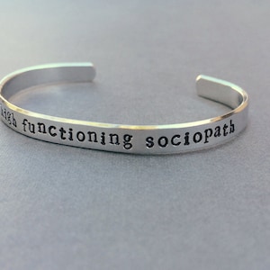 May include: A silver cuff bracelet with the words "high functioning sociopath" stamped in a black, typewriter-style font. The bracelet is open-ended and sits on a gray surface.