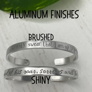 May include: Two silver bracelets with different finishes. The top bracelet is brushed and has the text "solemnly swear that I am up to no" engraved on it. The bottom bracelet is shiny and has the text "end of his roar, Sorrows will be" engraved on it.