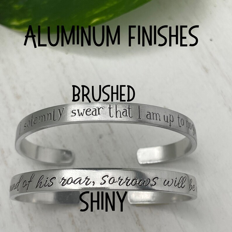 May include: Two silver bracelets with different finishes. The top bracelet is brushed and has the text "solemnly swear that I am up to no" engraved on it. The bottom bracelet is shiny and has the text "end of his roar, Sorrows will be" engraved on it.
