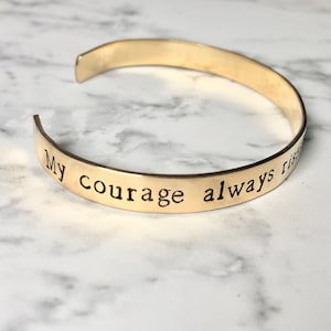 Könnte beinhalten: Goldfarbenes Metallarmband mit der Aufschrift "My courage always rises".