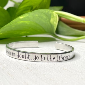 May include: A silver-colored metal cuff bracelet with an open design. The exterior surface of the bracelet is engraved with the black text "when in doubt, go to the library" in a simple, clean font. The bracelet has a smooth, polished finish.