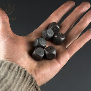 Può includere: Sei dadi grigio scuro con numeri bianchi sono tenuti in una mano aperta. I dadi sono di forma quadrata e sembrano essere di metallo. I numeri sono chiaramente visibili su ogni dado e la mano è davanti a uno sfondo scuro.