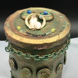 Peut inclure: Une boîte cylindrique verte et dorée avec un couvercle décoratif. Le couvercle est orné d'un motif de feuille d'or, de deux boutons bleus et d'une chaîne dorée. La boîte est décorée d'une chaîne, de boutons et d'une peinture verte et dorée.
