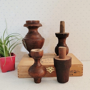 Könnte beinhalten: Vier antike Holzmöbelfüße in verschiedenen Formen und Größen, die satte Brauntöne aufweisen. Links steht ein kleiner rosa Topf mit einer grünen Pflanze. Die Beine sind auf einer Holzkiste mit goldfarbener Hardware angeordnet.