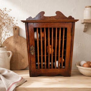 Op de afbeelding: Een rechthoekige houten eierkast met een latjesdeur aan de voorzijde, voorzien van een decoratief metalen schildje rond het sleutelgat. De kast heeft een gebogen bovenkant en bevat meerdere bruine en witte eieren op interne planken.