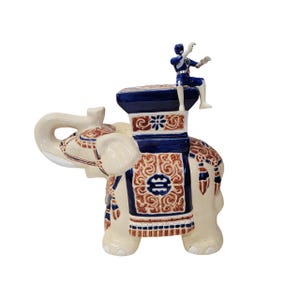 Könnte beinhalten: Eine Keramik-Elefantenfigur mit einem blau-braunen dekorativen Sattel und einer kleinen Figur oben. Der Elefant ist cremefarben mit detaillierten Mustern. Die kleine Figur trägt ein blau-weißes Outfit.