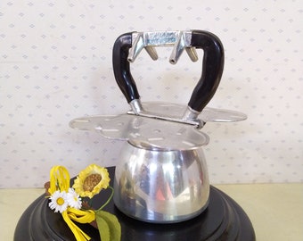 May include: A silver coffee maker with black handles sits on a black wooden base. The coffee maker has a unique design with a flower-shaped base and a round top. The coffee maker is surrounded by a yellow flower and green leaves.