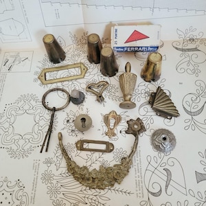 Puede incluir: Una colección de herrajes para muebles de latón y metal que incluye tiradores de cajón, cubiertas para bocallaves, remates decorativos y una pequeña caja con la etiqueta 25 PUNTINE FERRARI. Los artículos presentan formas variadas, incluyendo diseños rectangulares, circulares y florales.