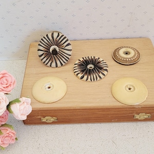 May include: Five circular decorative objects arranged on a rectangular wooden box. Two objects feature a dark brown radial sunburst pattern on a cream base, one has a brown dotted border, and two are plain cream-coloured discs with central holes.