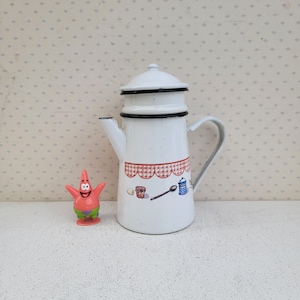Könnte beinhalten: Weiße Emaille-Kaffeekanne mit schwarzem Rand und rot-weißem Karomuster. Die Kanne hat einen Griff und einen Ausguss. Eine kleine, rosa Patrick Star-Figur steht neben der Kanne.