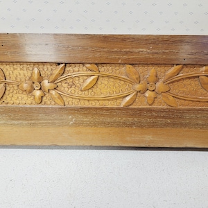 Può includere: Una sezione di modanatura decorativa in legno con motivi floreali e di foglie intagliati. La modanatura è di colore marrone chiaro con un bordo marrone più scuro. Il motivo floreale è centrato con foglie su entrambi i lati.