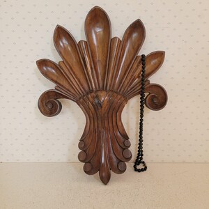 Puede incluir: Un elemento decorativo de pared de madera tallada con forma de abanico, con detalles intrincados y un color marrón intenso. Un collar de cuentas negras cuelga de un lado. El fondo es una pared beige claro con un patrón sutil.