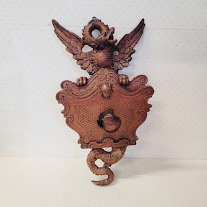 May include: A carved wooden wall hanging featuring a winged creature with a shield-shaped body and a serpent-like tail. The piece is made of dark brown wood and has intricate details, including carved feathers and a face on the shield.