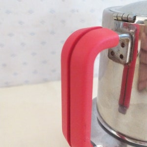 May include: A stainless steel coffee pot with a red handle. The pot has a hinged lid and a round base.