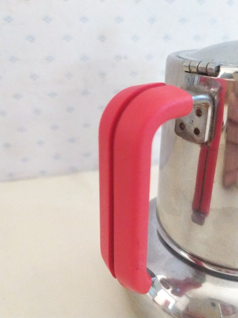 May include: A stainless steel coffee pot with a red handle. The pot has a hinged lid and a round base.