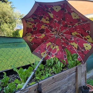 May include: An open umbrella with a floral pattern in red, yellow, and brown shades, positioned over a wooden planter box filled with leafy green plants. The umbrella's handle is silver, and the background includes a green fence and foliage.