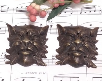 May include: Two antique bronze decorative door or cabinet pulls in the shape of a snarling, toothy, furry creature's face. The pulls are on a white sheet of music with a green leafy branch with red and white berries.