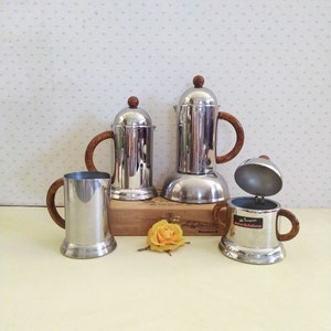 Peut inclure: Trois cafetières en argent avec des poignées en bois. Les cafetières sont sur une boîte en bois avec le texte "La Nuova  Numero 4" imprimé dessus. Une fleur jaune est devant la boîte.