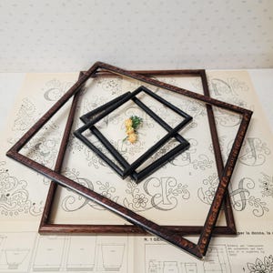 May include: A set of nested square picture frames including two large dark brown wooden frames with a textured finish and two smaller black frames. A small cluster of three artificial yellow flowers is placed in the centre of the frames.