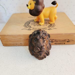Può includere: Una scultura dettagliata, in stile antico, di una testa di leone marrone è il punto focale, con una statuina di leone giallo e marrone sullo sfondo. La testa del leone è intagliata in modo intricato, mostrando una criniera e tratti del viso regali. Una scatola di legno con testo è dietro la statuina del leone.