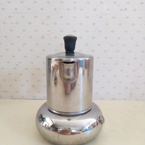 May include: A silver-colored stovetop espresso maker with a black knob on the lid. The maker is sitting on a white surface with a patterned background.