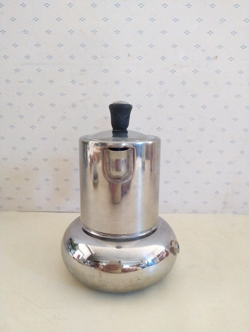 May include: A silver-colored stovetop espresso maker with a black knob on the lid. The maker is sitting on a white surface with a patterned background.