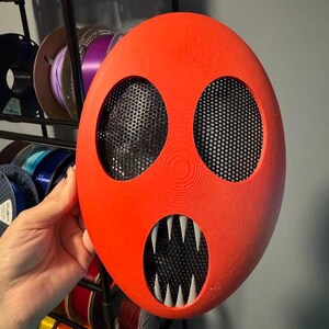 Pu&ograve; includere: Una maschera rosso vivo con due grandi fori per gli occhi e una bocca con affilati denti bianchi. Le aperture degli occhi e della bocca sono coperte da una rete nera. La maschera &egrave; tenuta davanti a un espositore di bobine colorate.