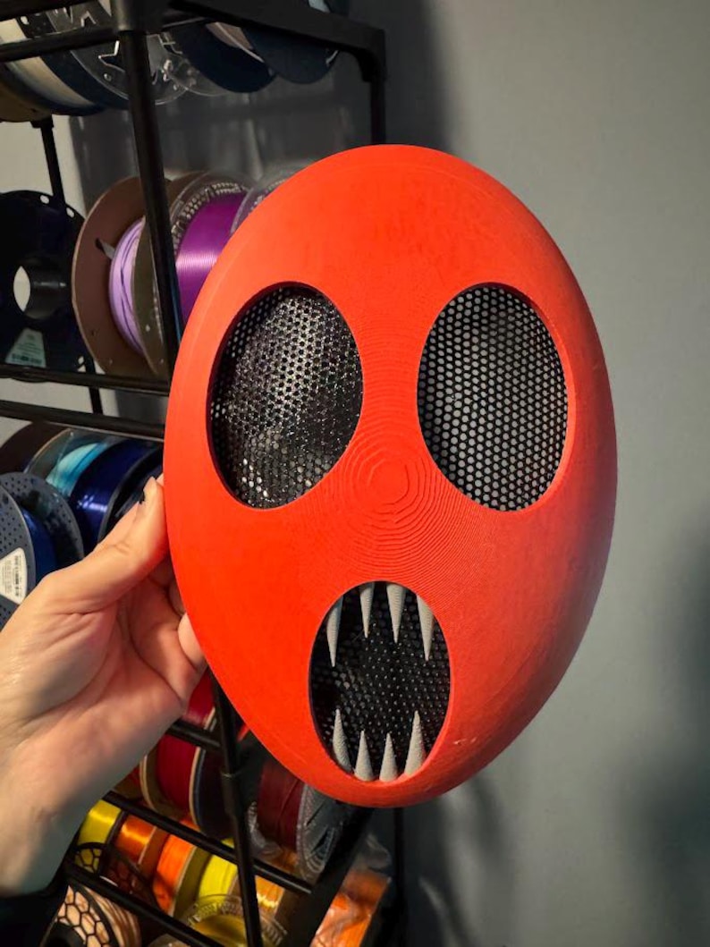 Pu&ograve; includere: Una maschera rosso vivo con due grandi fori per gli occhi e una bocca con affilati denti bianchi. Le aperture degli occhi e della bocca sono coperte da una rete nera. La maschera &egrave; tenuta davanti a un espositore di bobine colorate.