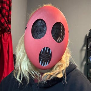 Pu&ograve; includere: Una maschera rossa con due grandi aperture circolari per gli occhi e una bocca con denti bianchi e affilati. Le aperture degli occhi e della bocca sono coperte da una rete nera. La maschera &egrave; indossata sul viso.