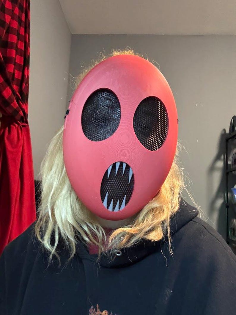 Pu&ograve; includere: Una maschera rossa con due grandi aperture circolari per gli occhi e una bocca con denti bianchi e affilati. Le aperture degli occhi e della bocca sono coperte da una rete nera. La maschera &egrave; indossata sul viso.