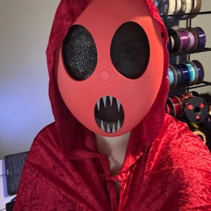 May include: A red hooded cloak and mask set. The mask is a solid red with large, black mesh eye openings and a toothy grin. The cloak is a deep red velvet material.