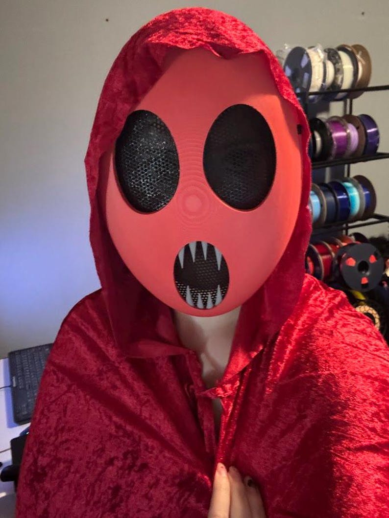 Pu&ograve; includere: Un set di maschera e mantello con cappuccio rossi. La maschera &egrave; di un rosso pieno con grandi aperture per gli occhi in rete nera e una bocca sorridente. Il mantello &egrave; in velluto rosso scuro.