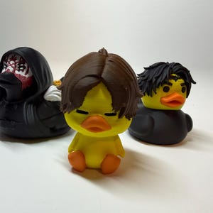 May include: Three rubber ducks featuring distinct designs. One is black with a hooded cloak and red facial markings. The center duck is yellow with brown hair and closed eyes. The third is yellow with black hair and a black body.