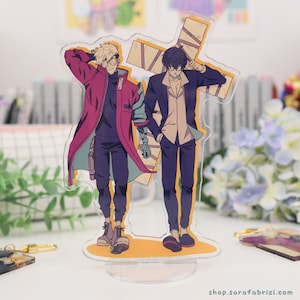 May include: Clear acrylic stand featuring two anime-style male figures. One figure wears a red coat, the other a dark suit. The stand has a yellow base and a cross-shaped design behind the figures. The website shop.sarafabrizi.com is printed at the bottom.