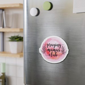 May include: A round magnet with the text "praying mamas club" in black script, surrounded by a watercolor-style pink and orange background and floral accents. The magnet is affixed to a silver refrigerator door.