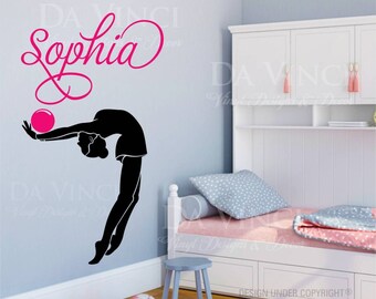 May include: A black silhouette of a gymnast holding a pink ball, with the name "Sophia" written in pink cursive above. The image is on a light blue wall with a white shelf unit and a pink and white bed in the background.