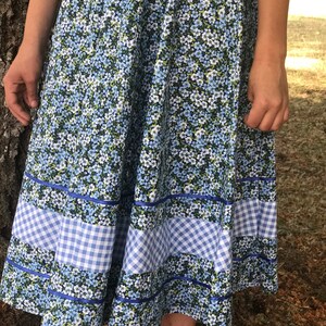 Puede incluir: Una falda floral azul y blanca con ribete de vichy. La falda es de largo hasta la rodilla y tiene una silueta completa y fluida.