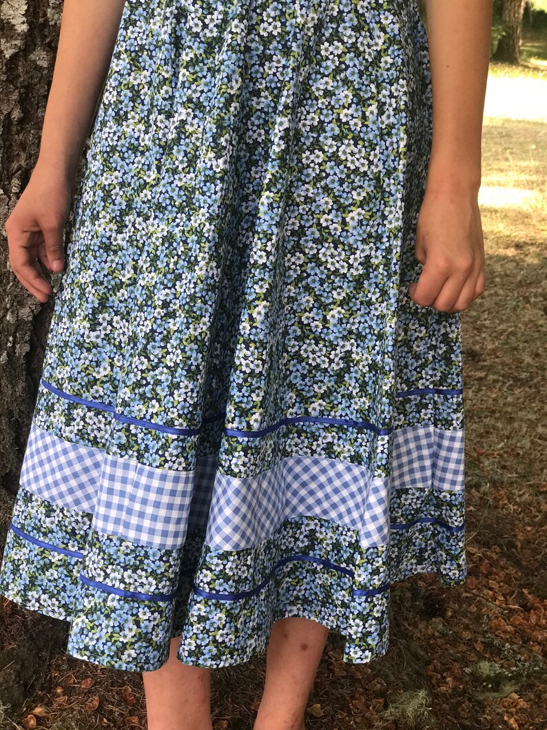 Puede incluir: Una falda floral azul y blanca con ribete de vichy. La falda es de largo hasta la rodilla y tiene una silueta completa y fluida.
