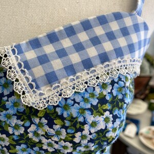 Puede incluir: Una tela de vichy azul y blanca con ribete de encaje blanco, superpuesta sobre una tela floral azul. La tela es probablemente para un vestido u otra prenda.