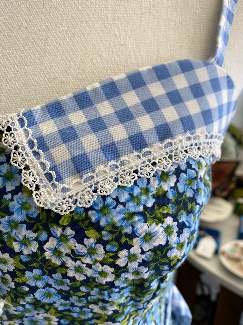 Puede incluir: Una tela de vichy azul y blanca con ribete de encaje blanco, superpuesta sobre una tela floral azul. La tela es probablemente para un vestido u otra prenda.