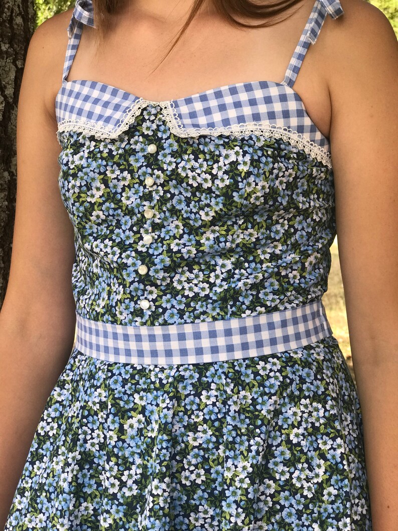 Puede incluir: Un vestido de verano azul y blanco con estampado de vichy y flores con ribete de encaje blanco y botones de perla. El vestido tiene un corpi&ntilde;o ajustado y una falda completa.