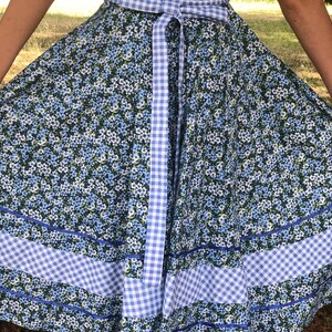 Puede incluir: Un vestido floral azul y blanco con una cinta de cuadros atada a la cintura. El vestido tiene una falda completa con ribete azul y ribete de cuadros.
