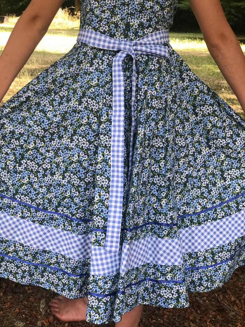 Puede incluir: Un vestido floral azul y blanco con una cinta de cuadros atada a la cintura. El vestido tiene una falda completa con ribete azul y ribete de cuadros.