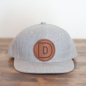 May include: A gray and brown baseball cap with a leather patch featuring the letter "D" in a circle.