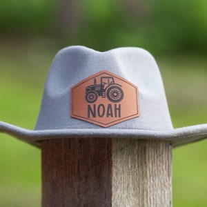 May include: A gray felt cowboy hat with a brown leather patch featuring a tractor and the name "NOAH".