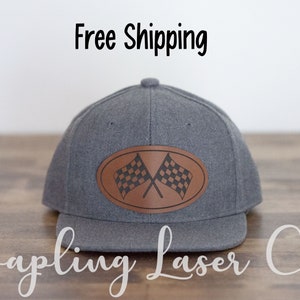 May include: Gray baseball cap with a brown leather patch featuring a checkered racing flag design. The text "Sapling Laser Co." is written below the cap.