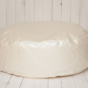 May include: A round, cream-colored beanbag chair with a smooth, satin-like surface. The beanbag is sitting on a white wooden floor.